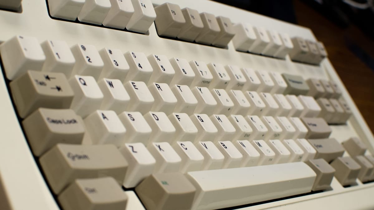 Close-up, angled view of an off-white mechanical keyboard with tall keycaps, black legends, and a classic layout, emphasizing the sculpted keys and tactile, old-school design.