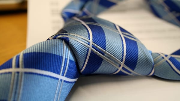 A close-up of a blue plaid necktie, focused on the knot with its textured fabric. A blurred sheet of paper and part of a keyboard sit in the background on a desk.