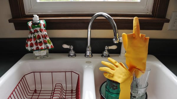 Yellow dishwashing gloves in a sink are propped on stacked glasses and a red pot, with one glove positioned to look like it’s giving the middle finger. A dish rack and soap sit nearby.